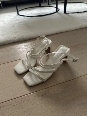 Vita klackskor  - Stilrena vita klackskor med fyrkantig tå, tunna remmar och justerbart spänne runt ankeln. Blockklack ger både höjd och stabilitet. Perfekta för dig som vill ha en clean och modern look till sommaren eller studenten. Använda en gång