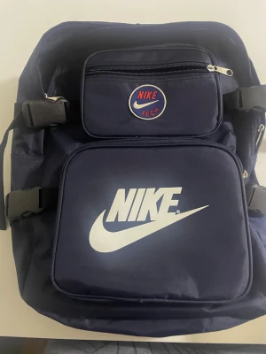 Mörkblå Nike Tech ryggsäck - Snygg mörkblå ryggsäck från Nike med stort vitt logotryck på framsidan och texten 'JUST DO IT.' på axelremmarna. Ryggsäcken har flera fack med dragkedja och justerbara remmar. Perfekt för skolan eller träningen.
