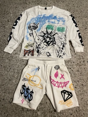 Vit långärmad tröja  - Vit långärmad topp med stort tryck av Frihetsgudinnan och New York-motiv framtill. Färgstänk i blått och svart samt grafiska detaljer på ärmarna. Rund halsringning och avslappnad passform, perfekt för en streetwear-look.