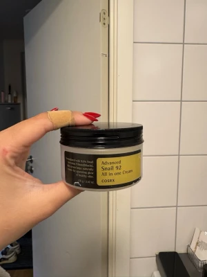 COSRX Advanced Snail 92 All in one Cream - En burk med COSRX Advanced Snail 92 All in one Cream. Krämen kommer i en rund, svart plastburk med guldfärgad etikett. Innehåller snigelsekretfiltrat som återfuktar och stärker huden. Perfekt för dig som vill boosta din hudvårdsrutin.