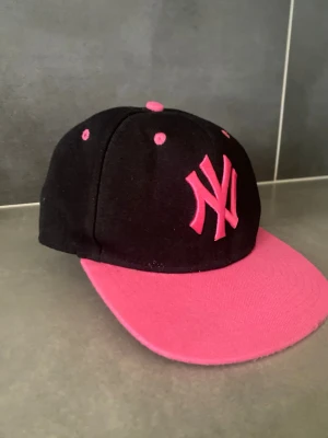 Svart och rosa New York Yankees keps - Säljer en snygg svart keps från New Era med rosa detaljer och broderad New York Yankees-logga framtill. Kepsen har platt skärm i rosa och matchande rosa knappar och ventilationshål. Perfekt för dig som vill sticka ut med en sportig och trendig stil.