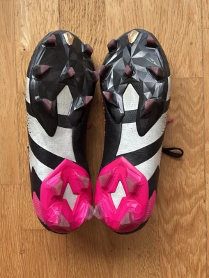 Adidas Predator svart/vit/rosa - Säljer ett par Adidas Predator fotbollsskor i svart, vitt och rosa. Skorna har en modern design med mönstrad ovandel, snörning och rosa detaljer på sulan. Perfekta för dig som vill ha grym kontroll på planen och sticka ut med snygg färgkombination.