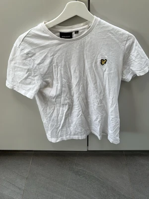 Vit t-shirt från Lyle & Scott - Klassisk vit t-shirt från Lyle & Scott med rund hals och korta ärmar. T-shirten har den ikoniska broderade loggan med gul örn på bröstet. Tillverkad i mjuk bomull för en skön känsla och enkel stil. Finns en lite defekt på armen som ni kan se på bild 3, annars är allt fint. Storlek xs