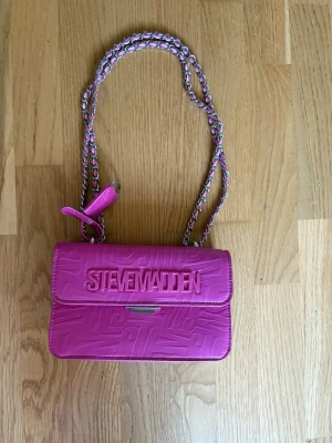 Rosa axelväska från Steve Madden - Cool rosa axelväska från Steve Madden med präglad logga på framsidan. Väskan har en kedjerem med inslag av rosa läder och ett dekorativt hänglås. Tillverkad i konstläder med stilren, rektangulär form och magnetlås.