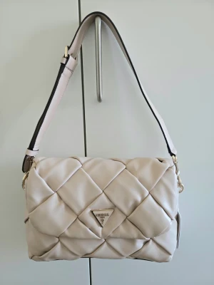 Beige axelväska från Guess - Beige axelväska från Guess (modell: Zaina). Mjukt läder och justerbar axelrem. Väskan har guldfärgade detaljer och en metallogo framtill. Jag fick väskan som en gåva för 2 år sedan, tycker om modellen men har inte använt den då jag sällan bär ljusa färger. Mycket bra skick: oanvänd, men fått små defekter av att ha legat under andra väskor i garderoben under en längre period. 