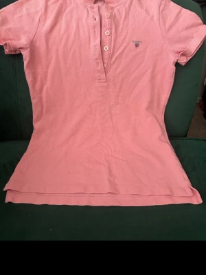 Rosa pikétröja från Gant - Säljer en klassisk rosa pikétröja från Gant med korta ärmar och knappar framtill. Tröjan har en liten broderad logga på bröstet och är tillverkad i mjuk bomull. Perfekt för en avslappnad och stilren look.