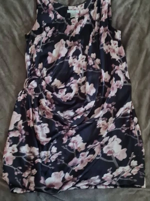 Blommig kortklänning från H&M - Supersöt blommig klänning från H&M i svart med rosa och vita blommor. Klänningen har ärmlös design, rund halsringning och en snygg detalj med ring i midjan. Perfekt för sommarens alla tillfällen!
