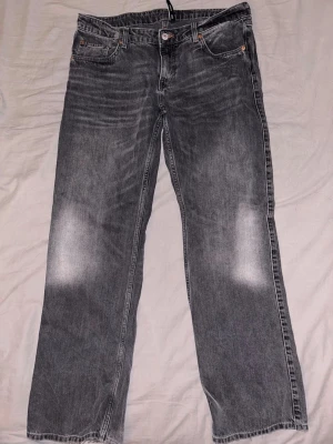 Lågmidjade raka jeans - Säljer ett par grå jeans med raka ben och snygga slitningar på knäna och vid bensluten. Jeansen har klassisk femficksdesign och en tvättad look som ger en cool vintagekänsla. Perfekta för en avslappnad streetstil. 32/32
