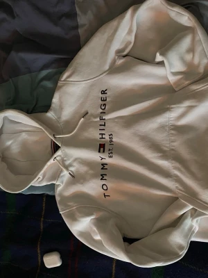 Vit hoodie från Tommy Hilfiger - Snygg vit hoodie från Tommy Hilfiger med logga och texten 'EST. 1985' på bröstet. Klassisk huva med dragsko och mjukt material som känns skönt mot huden. Perfekt för dig som gillar stilrena och tidlösa plagg.