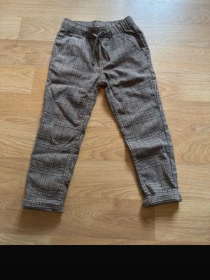 Rutiga byxor med dragsko - Snygga byxor i brunt och svart rutigt mönster med elastisk midja och dragsko. Byxorna har raka ben och sidofickor, perfekta för en avslappnad men trendig look. Materialet känns mjukt och bekvämt. Helt nya-oanvända!
