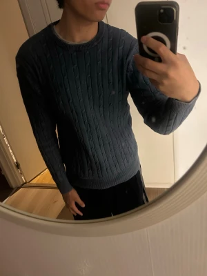 Blå kabelstickad tröja - Snygg mörkblå kabelstickad tröja med rund halsringning och ribbade muddar vid ärmslut och nederkant. Tröjan har en klassisk passform och är perfekt för lager-på-lager. Märket syns på etiketten i nacken.