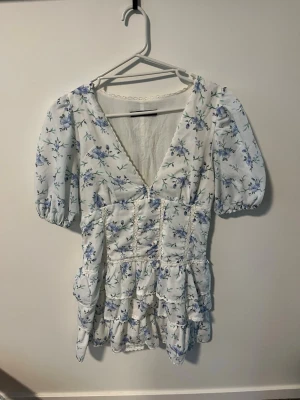 Blommig kortklänning med puffärm från Zara  - Söt kortklänning med blått blommönster på vit botten. Klänningen har v-ringning, puffärmar och volangdetaljer nedtill. Tillverkad i ett lätt och luftigt material, perfekt för varma dagar. Passformen är normal och klänningen har en romantisk och somrig vibe.