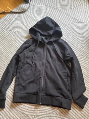 Svart hoodie med dragkedja från Hugo Boss - Snygg svart hoodie från Hugo Boss med dragkedja framtill och justerbar huva med snören. 