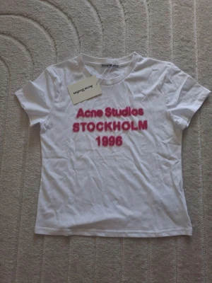 Vit Acne Studios t-shirt med tryck - Vit t-shirt från Acne Studios med rosa texttryck 'Acne Studios STOCKHOLM 1996' på bröstet. Klassisk rund halsringning och korta ärmar. Tillverkad i mjuk bomull för en skön känsla och stilren look.
