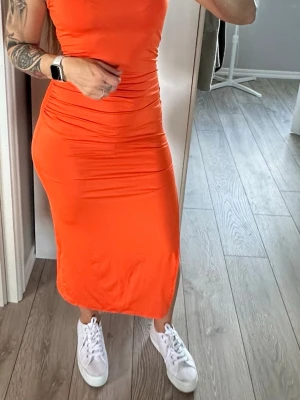 Orange tight midiklänning med slits - Snygg orange midiklänning i stretchigt material med figurnära passform. Klänningen har rynkade detaljer vid midjan och enslits på sidan. Strl S. Oanvänd. 