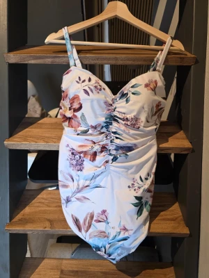 Blommig baddräkt med rynk framtill tummycontrol - Snygg baddräkt med vit bas och färgglatt blommönster i lila, rosa och grönt. Baddräkten har smala axelband och rynkdetalj framtill som ger en smickrande passform. Perfekt för strand eller poolhäng. Från next strl 42