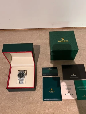 Role silverfärgad klocka med diamanter - Lyxig Role klocka i silver med svart urtavla och romerska siffror. Armbandet och boetten är täckta av gnistrande stenar för extra bling. Kommer i originalbox med certifikat och tillbehör. Perfekt för dig som vill sticka ut med en riktigt exklusiv accessoar.