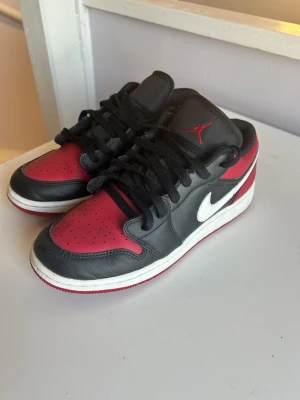 Air Jordan 1 Low svart/röd/vit - Snygga Air Jordan 1 Low sneakers i svart och rött läder med vit swoosh och röd yttersula. Klassisk siluett med svart snörning och röd Jumpman-logga på plösen. Perfekta för dig som gillar streetwear och ikoniska sneakers.
