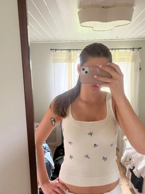 Vit stickad blommig topp - Supersöt vit stickad topp med breda axelband och små broderade blommor i lila och gult. Perfekt till sommaren och lätt att matcha med jeans eller kjol. Stretchigt material och croppad passform för en trendig look.