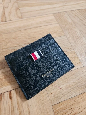 Svart kortfodral från Thom Browne - Snyggt svart kortfodral i präglat läder från Thom Browne. Fodralet har flera kortfack och en ikonisk tygdetalj i rött, vitt och blått framtill. Perfekt för dig som vill ha en stilren och exklusiv accessoar i fickan.