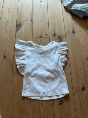 Vit topp med volangärmar - Superfin vit topp med breda volangärmar som ger en trendig och söt look. Toppen har rund halsringning och är tillverkad i ett mjukt, ribbat material som känns skönt mot huden. Perfekt att styla med jeans eller kjol för en fräsch outfit.
