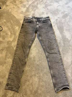 Grå slim fit jeans med knappar - Säljer ett par grå slim fit jeans med snygga knappar på fickan och klassiska femficksdetaljer. Jeansen har en tvättad look och raka ben, perfekt för dig som gillar en avslappnad stil. Materialet är denim och färgen är grå.