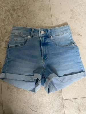 Ljusblå denimshorts med uppvik - Snygga ljusblå denimshorts med mid Waist och uppvikta benslut. Klassisk femficksmodell med knapp och dragkedja framtill. Perfekta för varma dagar och enkla att matcha med olika toppar.