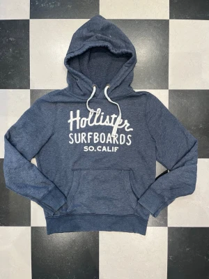 Blå Hollister hoodie - Snygg blå vintage hoodie från Hollister med vit text 'Hollister Surfboards SO. CALIF' på bröstet. Klassisk känguruficka framtill och vita dragsnören i huvan. Tröjan har små hål på framsidan som knappt syns när man bär den. Mjuk bomullsblandning och avslappnad passform, perfekt för chill dagar.