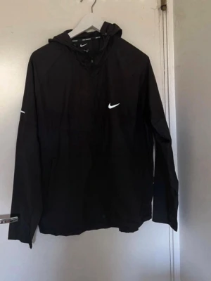 Svart Nike vindjacka med huva - Snygg svart vindjacka från Nike med vit logga på bröstet och huva. Jackan har dragkedja framtill och långa ärmar. Perfekt för blåsiga dagar och enkel att matcha med olika outfits.