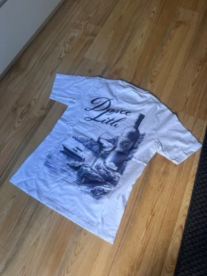 Vit t-shirt med vintagemotiv - Vit t-shirt med stort tryck på ryggen som föreställer en vinflaska, vinglas och en båt vid vattnet. Texten 'Dolce Lith' syns i motivet. Klassisk passform och korta ärmar, perfekt för en chill och avslappnad stil.