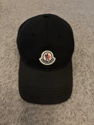 Svart keps från Moncler - Snygg svart keps från Moncler med klassisk böjd skärm och broderad Moncler-logga framtill. Kepsen är i bomull och har ventilerande hål upptill. 