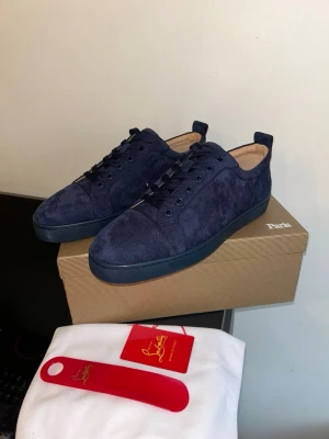 Mörkblå sneakers från Louboutin - Stilrena mörkblå sneakers från Christian Louboutin i mjuk mocka med matchande mörkblå sula och ton-i-ton snörning. Låg modell med diskret broderad logga på hälen och beige innersula. Perfekta för dig som gillar exklusiva detaljer och klassisk design.