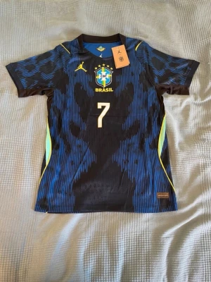 Brasilien Vinicius Jr matchtröja blå - Snygg blå och svart Brasilien matchtröja från Nike med nummer 7 och Vinicius Jr på ryggen. Tröjan har gula detaljer, CBF-logga och Nike-logga på bröstet samt moderna mönster. Perfekt för dig som älskar fotboll och vill stötta landslaget.