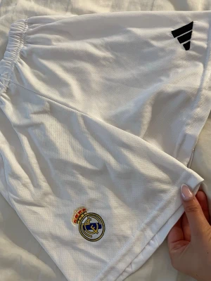 Vita Real Madrid shorts från Adidas - Säljer ett par vita sportshorts från Adidas med Real Madrid-logga broderad på ena benet och Adidas-logga i svart på andra benet. Shortsen har elastisk midja och är tillverkade i ett lätt, ventilerande material som passar perfekt till träning eller match.