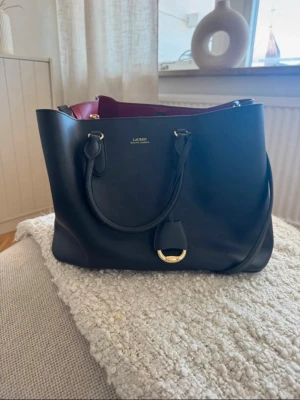 Svart handväska från Lauren Ralph Lauren - Stilren svart handväska i slätt läder från Lauren Ralph Lauren. Väskan har guldfärgade metalldetaljer, dubbla handtag och en snygg vinröd insida. Perfekt för dig som vill ha en klassisk och rymlig väska med exklusiv känsla.