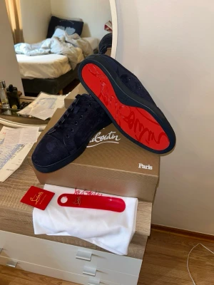 Mörkblåa Louboutin sneakers - Snygga mörkblåa sneakers från Christian Louboutin i mocka med klassisk röd sula och diskret broderad logga på hälen. Skorna har lågt skaft och svarta skosnören. Perfekta för dig som vill sticka ut med exklusiv stil.