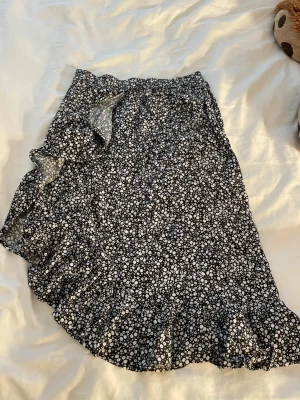 Svart blommig omlottkjol med volang - Säljer en svart omlottkjol med vita små blommor och volangkant. Kjolen har en feminin look med knyt i sidan och är perfekt för dig som gillar en söt och trendig stil. Passar till både sneakers och boots.