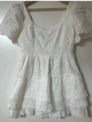 Meet me there klänning - Superfin vit kort klänning med broderade blommor och volangkant. Klänningen har puffiga korta ärmar och rundad halsringning. Perfekt till sommaren