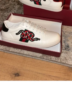 Gucci vita sneakers med orm - Snygga vita sneakers från Gucci med broderad röd och svart orm på sidan samt klassiska gröna och röda ränder. Skorna har svarta och röda detaljer på hälen och är tillverkade i läder. Perfekt för dig som vill sticka ut med en lyxig och trendig look.