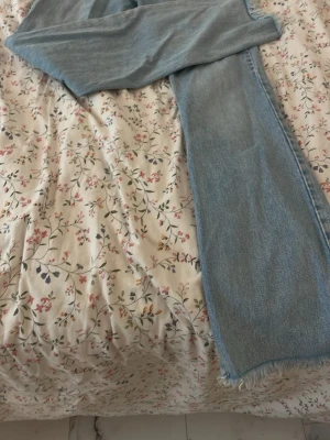 Ljusblå raka jeans med rå kant - Säljer ett par ljusblå jeans med rak passform och rå, fransad kant nertill. Jeansen har klassisk femficksdesign och är tillverkade i denim. Perfekta för en avslappnad och trendig look.