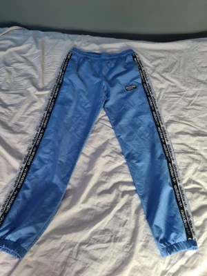 Blå adidas mjukisbyxor med logga - Snygga ljusblå mjukisbyxor från adidas med svarta och vita loggor längs sidorna. Byxorna har resår i midjan och vid bensluten samt en broderad adidas-logga på ena benet. Perfekta för en avslappnad och sportig stil.