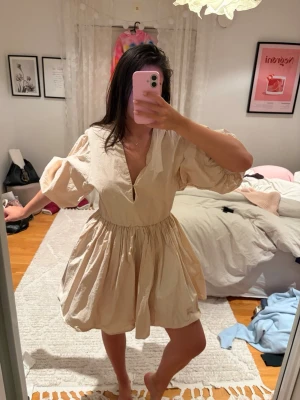 Beige kort klänning med puffärm - Säljer en beige kortklänning med v-ringning och voluminösa puffärmar. Klänningen har en markerad midja och ett luftigt, utställt kjolparti. Perfekt för dig som gillar en avslappnad men trendig stil.