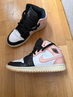 Air Jordan 1 Mid svart/rosa/vit - Snygga Air Jordan 1 Mid sneakers i svart, vitt och ljusrosa läder. Klassisk siluett med svart snörning, vit tåbox och rosa detaljer runt häl och Swoosh. Perfekta för dig som vill sticka ut med stilrena färgkombos och ikonisk design.