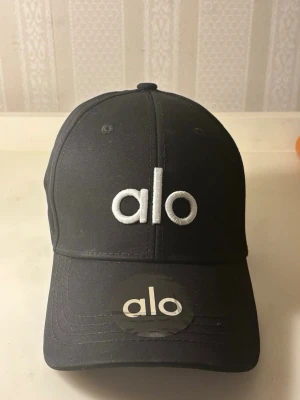 Svart keps från Alo med broderad logga - Snygg svart keps från Alo med böjd skärm och broderad vit logga framtill. Klassisk baseballmodell med justerbar baksida. Tillverkad i slitstarkt tyg som passar perfekt till streetwear eller sportiga outfits.