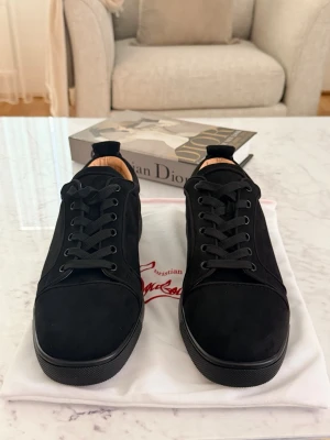 Svarta sneakers från Christian Louboutin - Snygga svarta sneakers från Christian Louboutin i mocka med klassisk röd sula och svarta skosnören. Låg modell med stilren design och diskreta detaljer. Perfekta för dig som vill ha en clean look med exklusiv känsla.