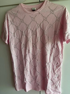 Rosa t-shirt med silverpaljetter - Säljer en ljusrosa t-shirt från river Island med korta ärmar. T-shirten har ett unikt mönster av silverfärgade diamanter över hela framsidan som ger en snygg och trendig look. Tillverkad i mjuk bomull för skön känsla och passform. Den är lite skrynklig men jag kan strycka den till den som köper. Tycker den är ganska stor och skulle passa en vuxen också.