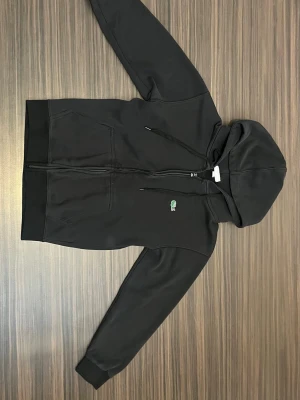 Zip hoodie från Lacoste - Ljus svart hoodie med dragkedja från Lacoste. Klassisk krokodil-logga på bröstet, två fickor framtill och justerbar huva med snören. Tillverkad i mjukt material 