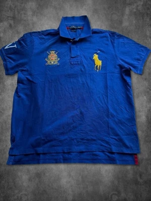 Blå piké från Polo Ralph Lauren XL - Snygg blå pikétröja från Polo Ralph Lauren i bomull, custom fit. Klassisk krage, kort ärm och broderad gul logga på bröstet samt emblem. Perfekt för en clean och sportig look.