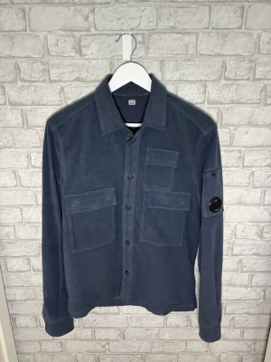 C.P. Company overshirt - Riktigt fet C.P. Company overshirt i mörkblå färg med klassiska lens-detaljen på armen. Mjukt mocka-liknande material med premiumkänsla och riktigt skön passform. Funkar perfekt både som overshirt och tunnare jacka nu till vår/sommaren.