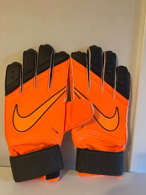 Nike orangea målvaktshandskar - Snygga målvaktshandskar från Nike i starkt orange med svarta detaljer och stor logga. Handskarna har breda kardborreband runt handleden och insidan är vit för bra grepp. Perfekta för dig som vill sticka ut på planen. Använd max tre gånger.